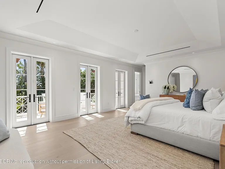This is a bright and spacious primary bedroom featuring white walls and light wood flooring. The room has access to outdoor balconies through double doors, enhancing the airy feel. A large, made bed dominates the space, complemented by a round mirror and minimal decor for a clean and modern look.