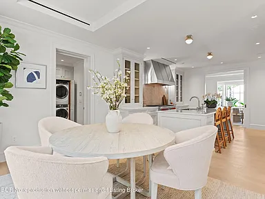 This is an interior shot of a bright and modern kitchen. A round dining table with upholstered chairs sits in the foreground, while the kitchen features white cabinetry, stainless steel appliances, and an island. The space exudes a clean, sophisticated, and inviting atmosphere.