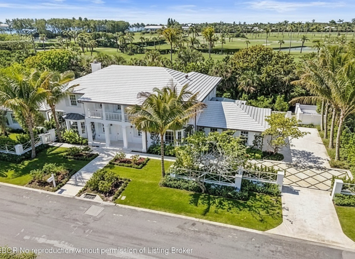 This is an aerial view of a luxurious, two-story home with a white tile roof situated on a lush green lot. The property showcases meticulously manicured landscaping, including well-defined garden beds, mature palm trees, and a manicured lawn. A long, patterned concrete driveway leads to an ornate gate, adding to the property's curb appeal.
