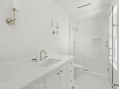 The image shows a bright, all-white bathroom. It features a vanity with white cabinets and a countertop, a sink, and brushed nickel fixtures. A glass-enclosed shower with white tile is present, along with a toilet, making it a functional and clean space.