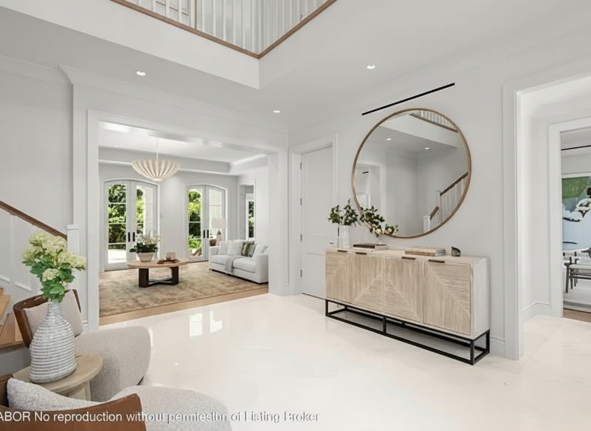 This is a well-lit interior shot of a hallway featuring crisp white walls and bright flooring. The space showcases a round mirror above a decorative cabinet, enhancing the entryway's appeal. The hallway leads to an open living area, creating an inviting and spacious feel.