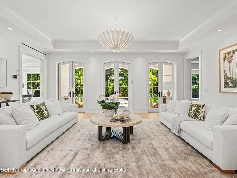 The living room features two white sofas with decorative pillows facing each other across a large area rug. A round, light-colored coffee table sits in the center. Large arched doorways and windows provide ample natural light, emphasizing the bright and airy atmosphere of the space. A unique chandelier hangs from the ceiling, adding a touch of modern elegance.