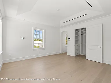 The image showcases a bright and spacious primary bedroom. It features light hardwood flooring, white walls, and a walk-in closet with white doors. A window provides natural light, and overall the design is modern and minimalist.
