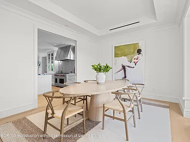 The image showcases an elegant dining room featuring a light wood oval dining table surrounded by stylish chairs with woven seats. A large abstract artwork adorns the wall, adding a touch of sophistication. The room is bright and airy, with white walls and light wood flooring creating a clean and modern aesthetic.