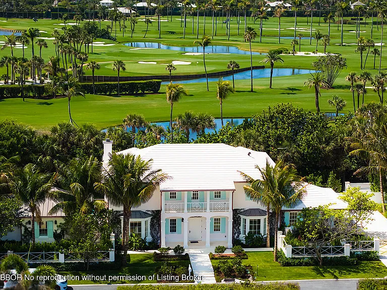 This aerial view showcases a beautiful white house with a well-manicured lawn, palm trees, and a pristine golf course in the background. The architectural style is elegant and refined, presenting a luxurious residential property. The setting suggests a high-end community focused on leisure and recreation.