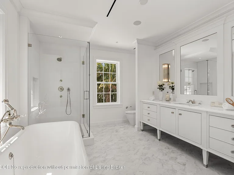 This is a bright and elegant primary bathroom featuring a freestanding bathtub with a silver faucet, separate glass-enclosed shower, and a double vanity with white cabinetry and light-colored countertop. The floor is covered in marble, and large mirrors are hung above the vanity. Overall, the bathroom exudes a luxurious and clean aesthetic.