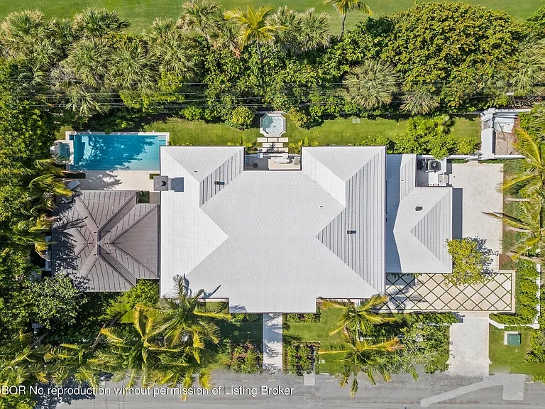 This aerial shot showcases an expansive property featuring a large house with a light-colored roof, a private swimming pool, and a spa. The surrounding landscape includes lush greenery and palm trees, enhancing the luxurious feel of the estate. The driveway has a decorative pattern that compliments the landscape design.