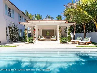 This image showcases a luxurious pool area of a residential property. The crystal-clear pool water leads to a covered patio area with lounge seating. The surrounding landscape features lush tropical plants, creating a private and serene backyard oasis.
