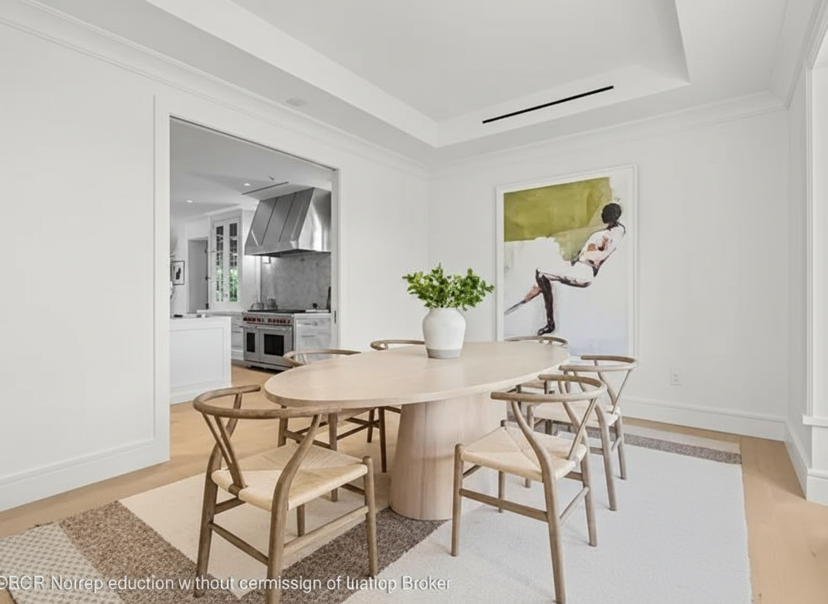 This is an interior shot of a dining room featuring a light wood oval dining table surrounded by six chairs with woven seats. The room has white walls and trim, and an adjacent kitchen is visible through a doorway. A large painting hangs on the wall, and there is a neutral-toned area rug under the table.