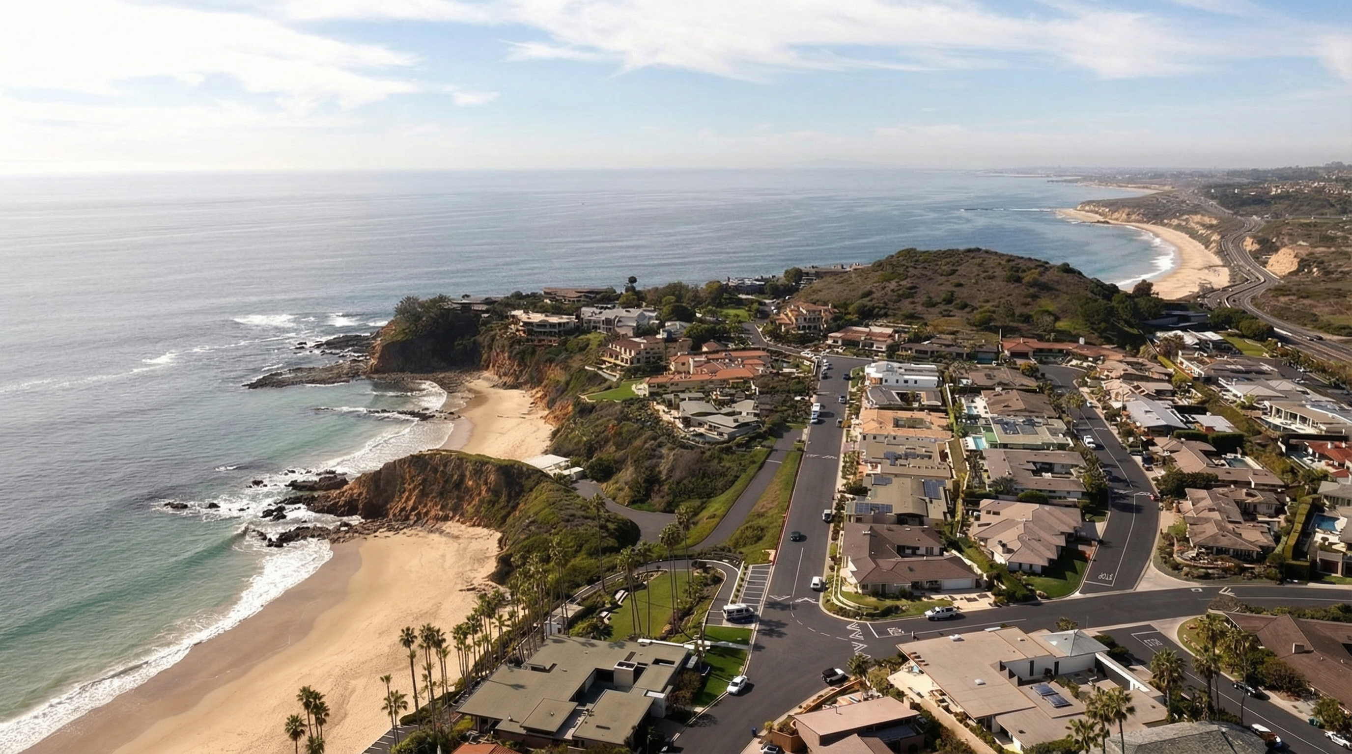 This aerial view showcases a coastal residential area featuring luxury homes with terracotta roofs, lush landscaping, and ocean views. The properties are ideally situated near sandy beaches and a highway, making this a desirable location. The image highlights the area's natural beauty and luxurious lifestyle.