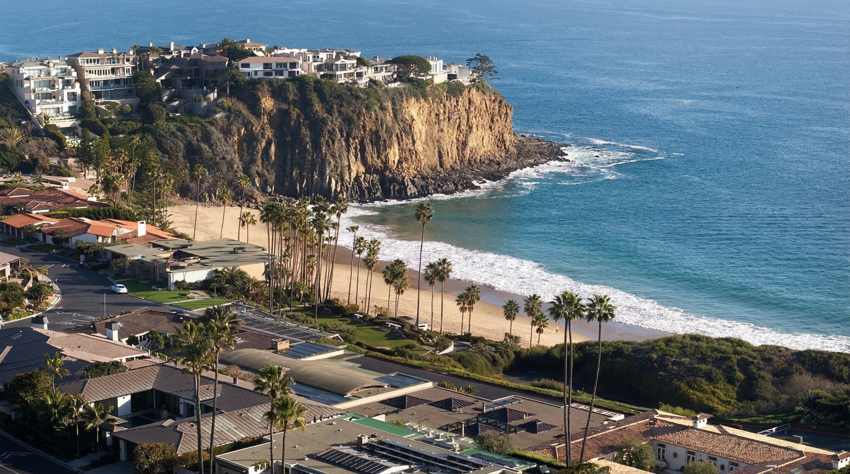 This aerial view showcases a luxurious coastal neighborhood with stunning oceanfront properties. The image features elegant homes perched atop a cliff, a sandy beach lined with palm trees, and the clear blue waters of the ocean. The scene conveys a sense of exclusivity and high-end living in a desirable location.