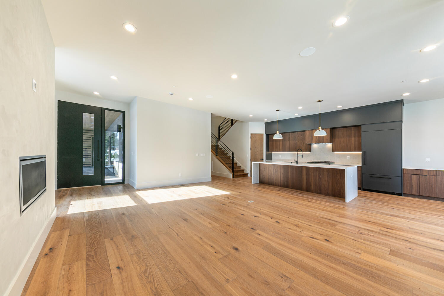 This is an interior shot showcasing a generously sized living room that seamlessly integrates with an open-concept kitchen. The living area features hardwood flooring, bright lighting, and a modern fireplace accent. The kitchen boasts a large island, wood cabinetry, and sleek appliances, creating a harmonious and contemporary living space, which makes it very appealing to potential homeowners.