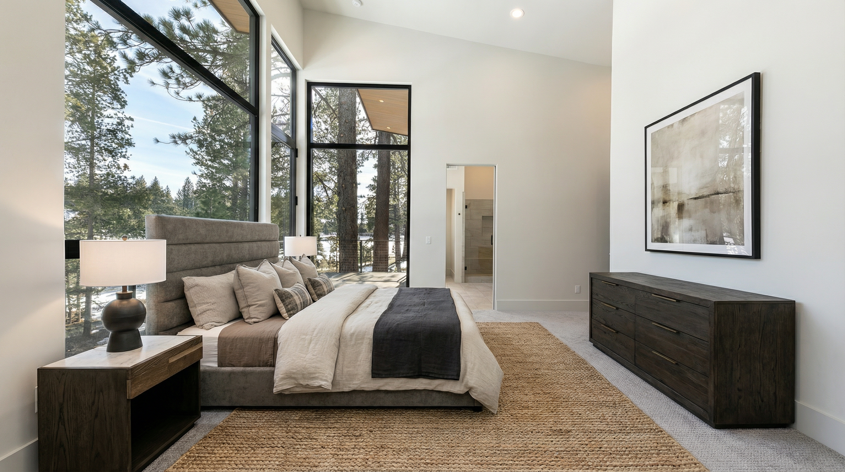 This is a primary bedroom featuring a large window with views of surrounding trees and water. The room is decorated in neutral tones with a grey upholstered bed, a woven rug, and a dark wood dresser offering ample storage. The bedroom has an ensuite bathroom, adding to the suite's appeal.