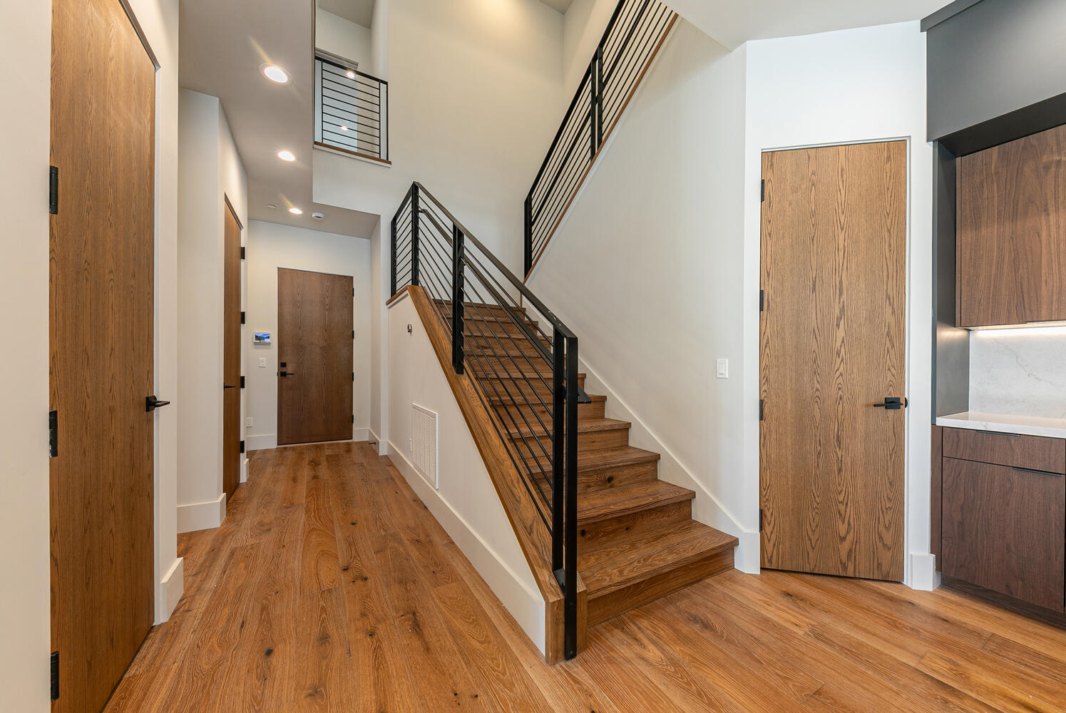 This interior shot showcases a well-lit hallway with wooden flooring and stairs. The staircase has modern black metal railings, leading to an upper level, and wood doors provide access to adjacent rooms. The scene presents a clean and contemporary aesthetic, highlighting the home's architectural details.