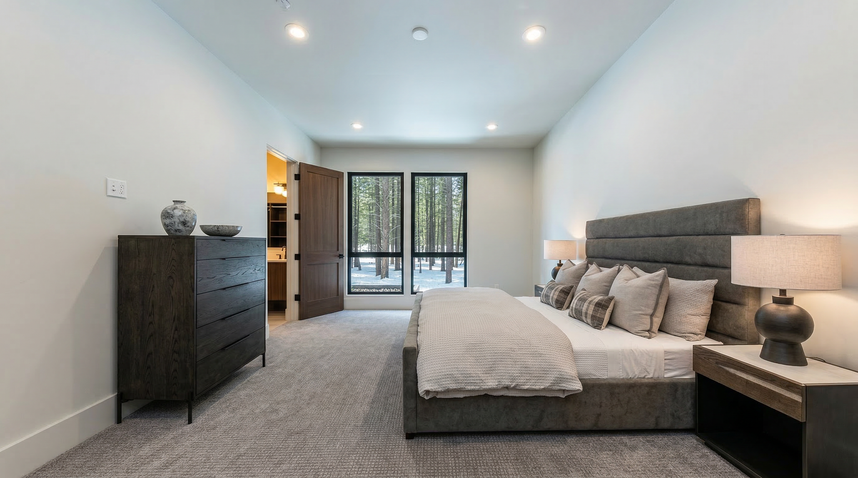 This is a well-lit primary bedroom featuring a gray upholstered bed with a large headboard, a dark wood dresser with decorative items on top, and a matching nightstand with a lamp. The neutral color palette and plush carpet create a serene and inviting atmosphere. Large windows offer natural light and a view of the outdoors.