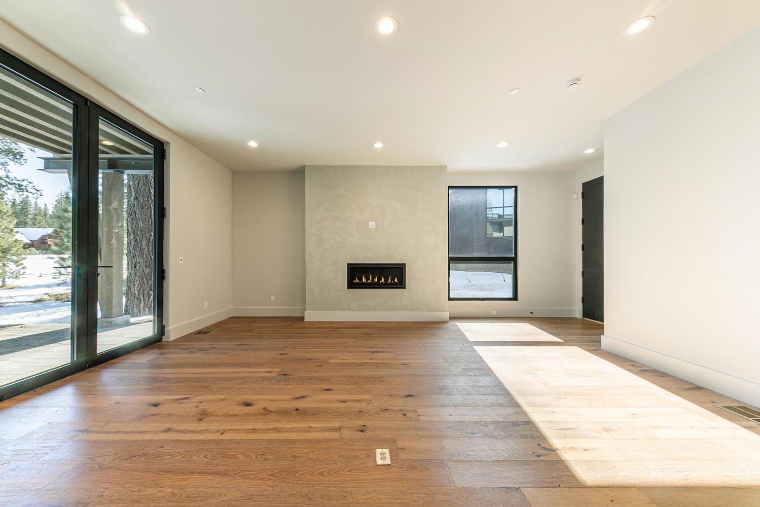 The image shows a spacious living room with hardwood flooring and a modern fireplace. Large windows and glass doors provide ample natural light. The room has a minimalist design with clean lines and neutral wall colors, creating an airy and inviting atmosphere.