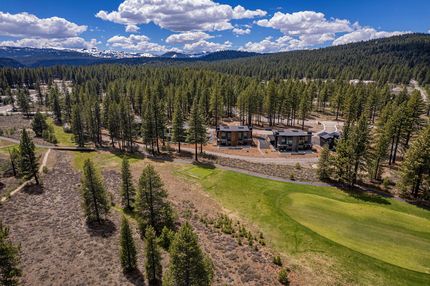 This aerial shot showcases a modern residential development nestled within a lush forest. Two contemporary homes with dark exteriors and clean lines are visible, set against a backdrop of tall evergreen trees and distant snow-capped mountains. A well-manicured green expanse, possibly a golf course or expansive lawn, adds to the property's appeal, highlighting the serene and private setting.