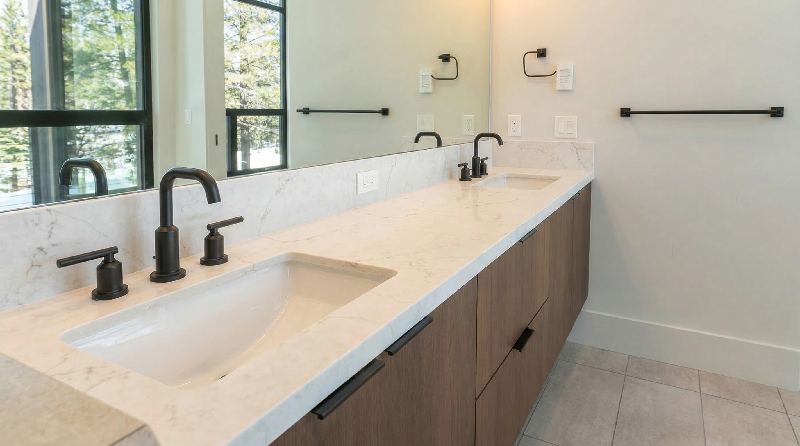 This is a well-lit primary bathroom featuring a double vanity with a white countertop and dark wood drawers and cabinets. The fixtures are a modern matte black, and a large mirror spans the length of the vanity. The overall design is clean and contemporary.
