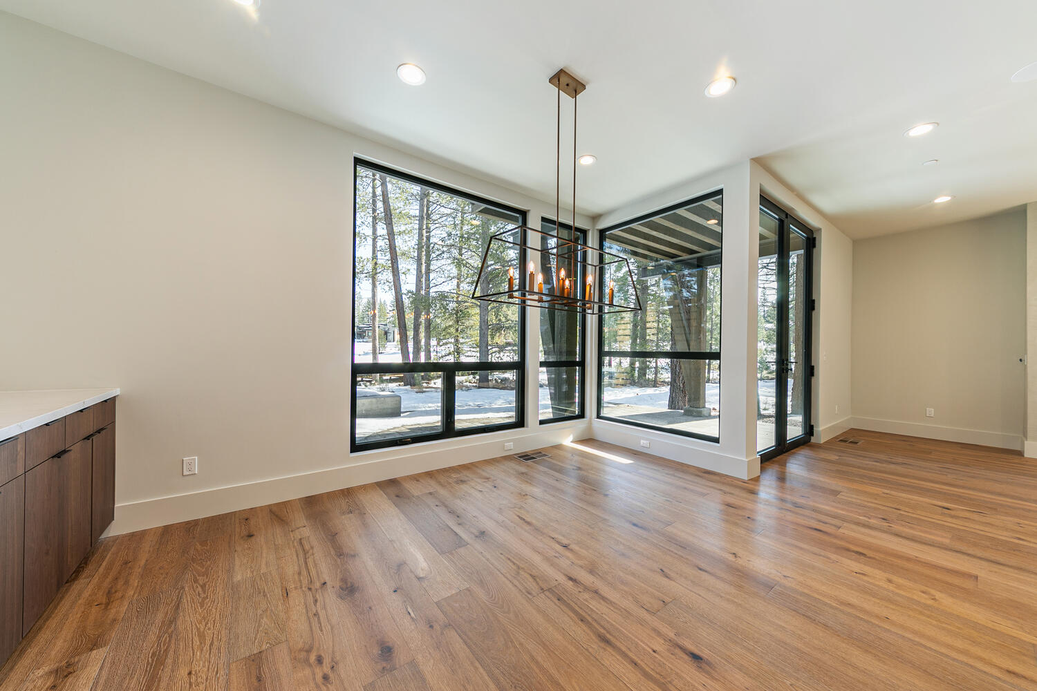 This is a dining room showcasing a modern design with hardwood floors and large windows that provide ample natural light and views of the surrounding landscape. A contemporary chandelier hangs above the dining area. The space is clean, open, and inviting, perfect for both everyday meals and formal gatherings.