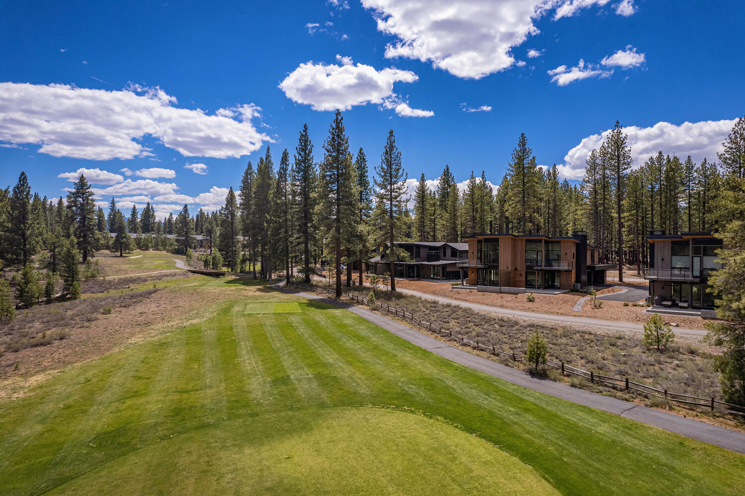 This aerial view showcases a modern property nestled on a golf course. The residence features contemporary architectural design with clean lines and a natural material palette complementing the surrounding trees. The meticulously maintained green lawn and asphalt path enhance the property's appeal.