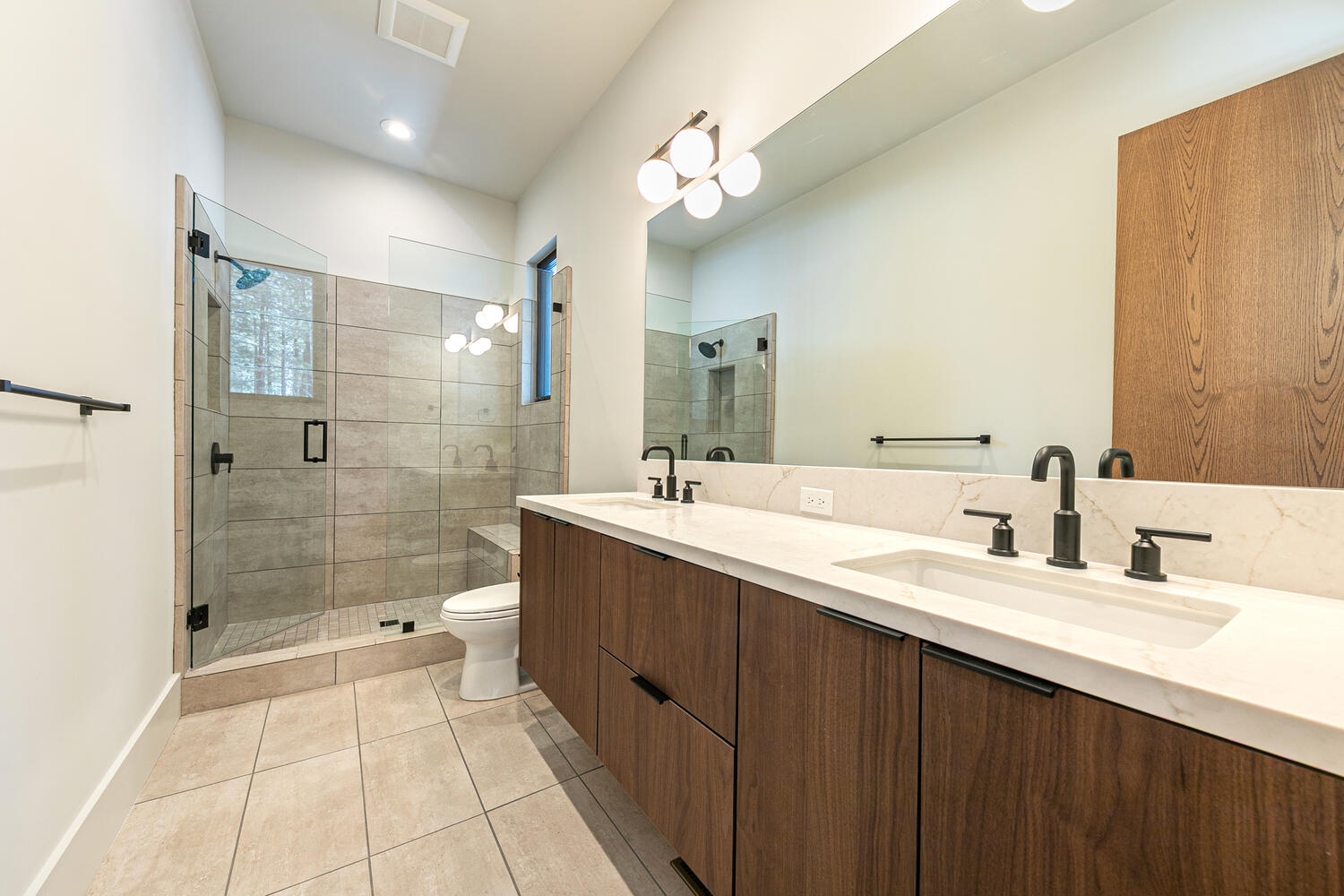 This is a contemporary primary bathroom featuring a double vanity with a white countertop and dark wood cabinetry. A large mirror spans the length of the vanity wall. A glass-enclosed shower is visible to the left, showcasing tiled walls and a built-in bench, and there's a toilet situated between the shower and vanity.