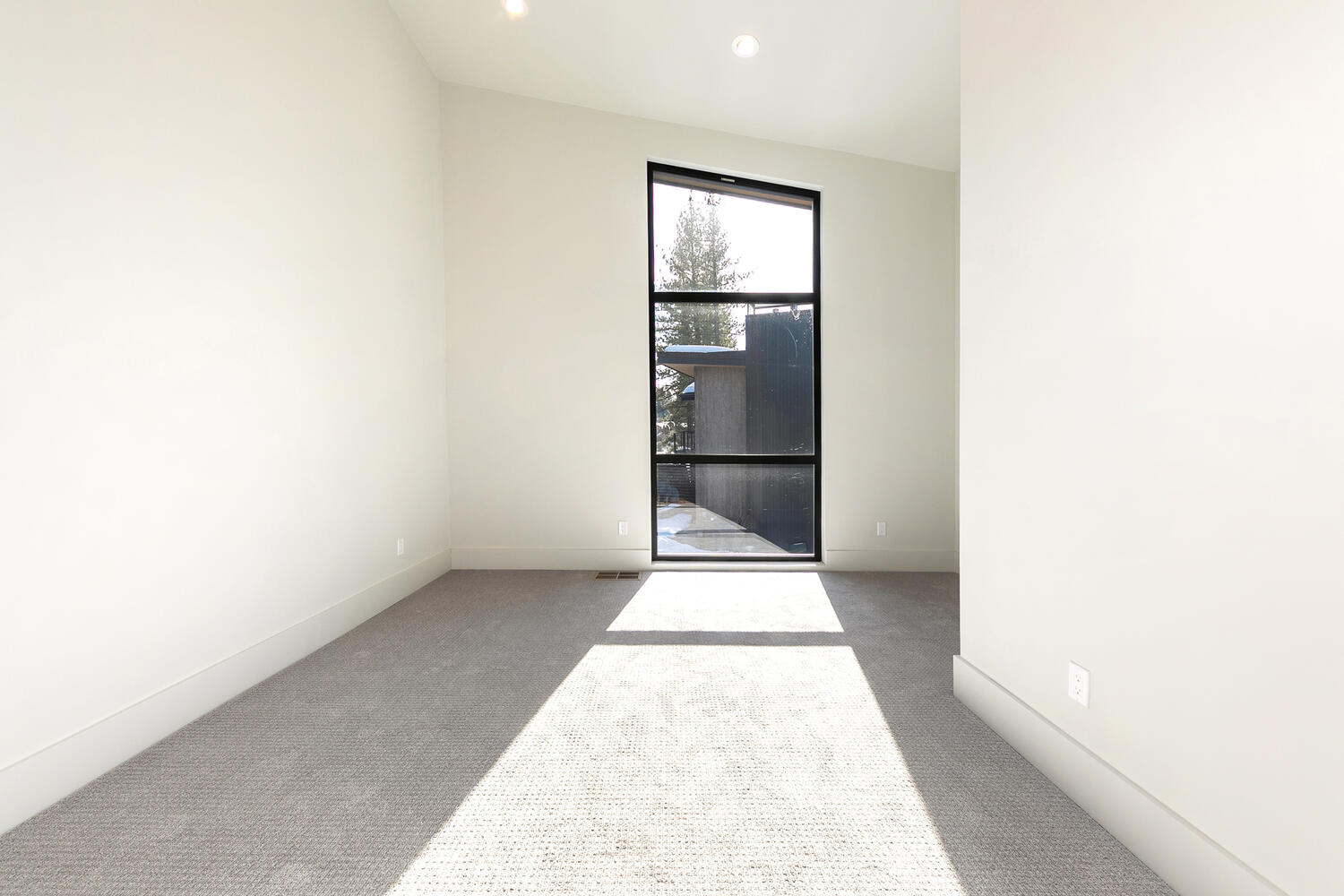 This is a bright, modern bedroom with neutral colors. The room features gray carpet, pale walls, and a tall window with a black frame offering natural light. The space is clean and minimalistic, ideal for a guest bedroom or a flexible living space.