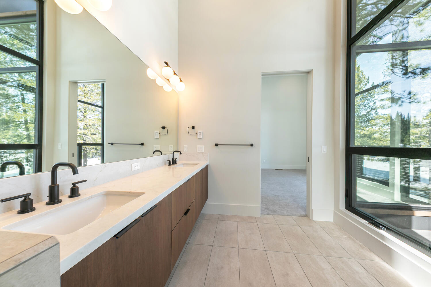 This is a well-lit primary bathroom featuring a double sink vanity with a white countertop and dark wood cabinetry. A large mirror spans the length of the vanity, reflecting the light from modern sconces and natural light from a grand window. The room has a clean, contemporary aesthetic with light-colored floor tiles and neutral walls, conveying a sense of spaciousness and luxury.