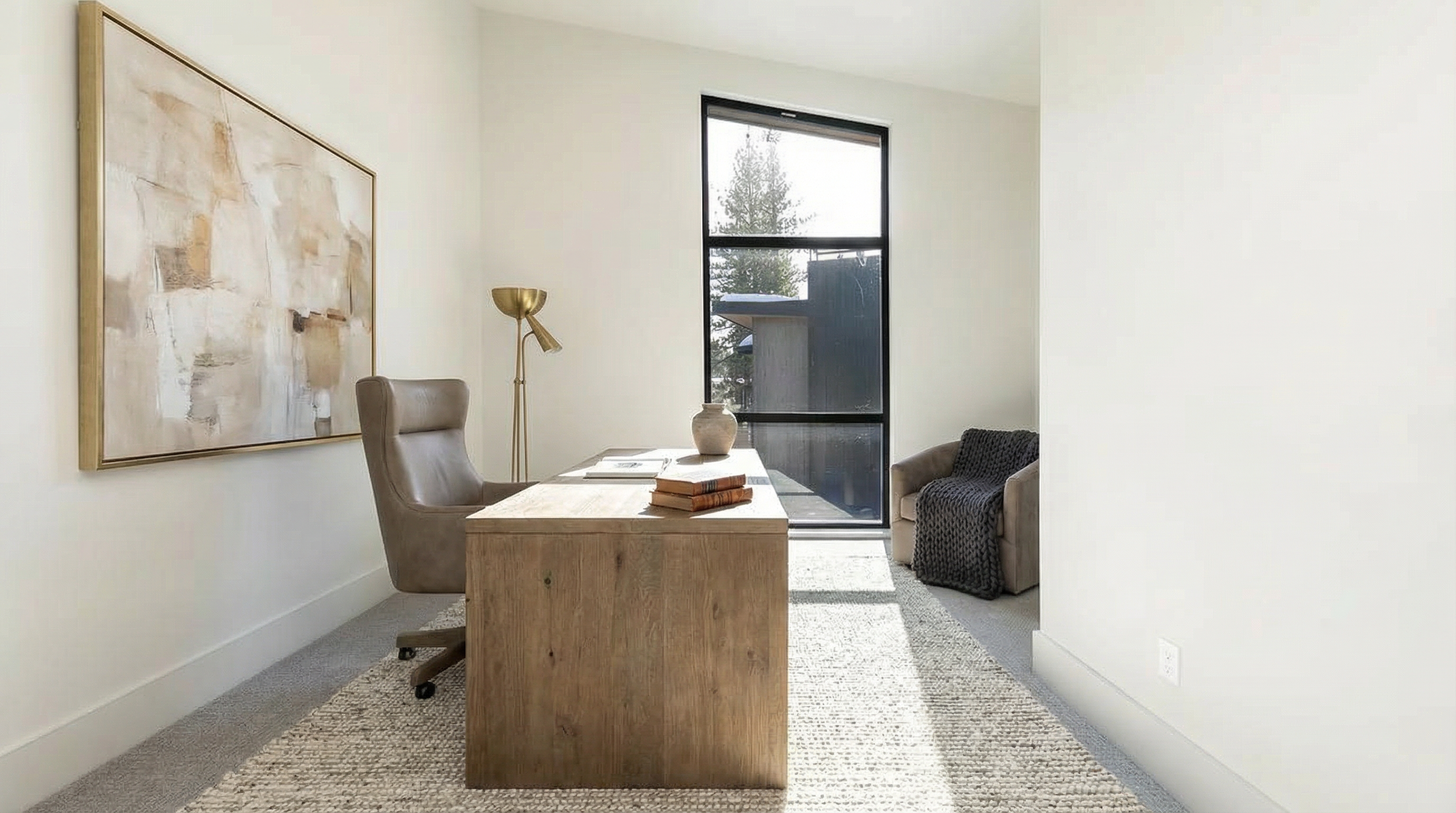 This is a well-lit home office featuring a wooden desk, a comfortable chair, and large abstract art. A tall window provides natural light and a view to the outdoors. A neutral color palette and a textured rug create a calm and sophisticated workspace.