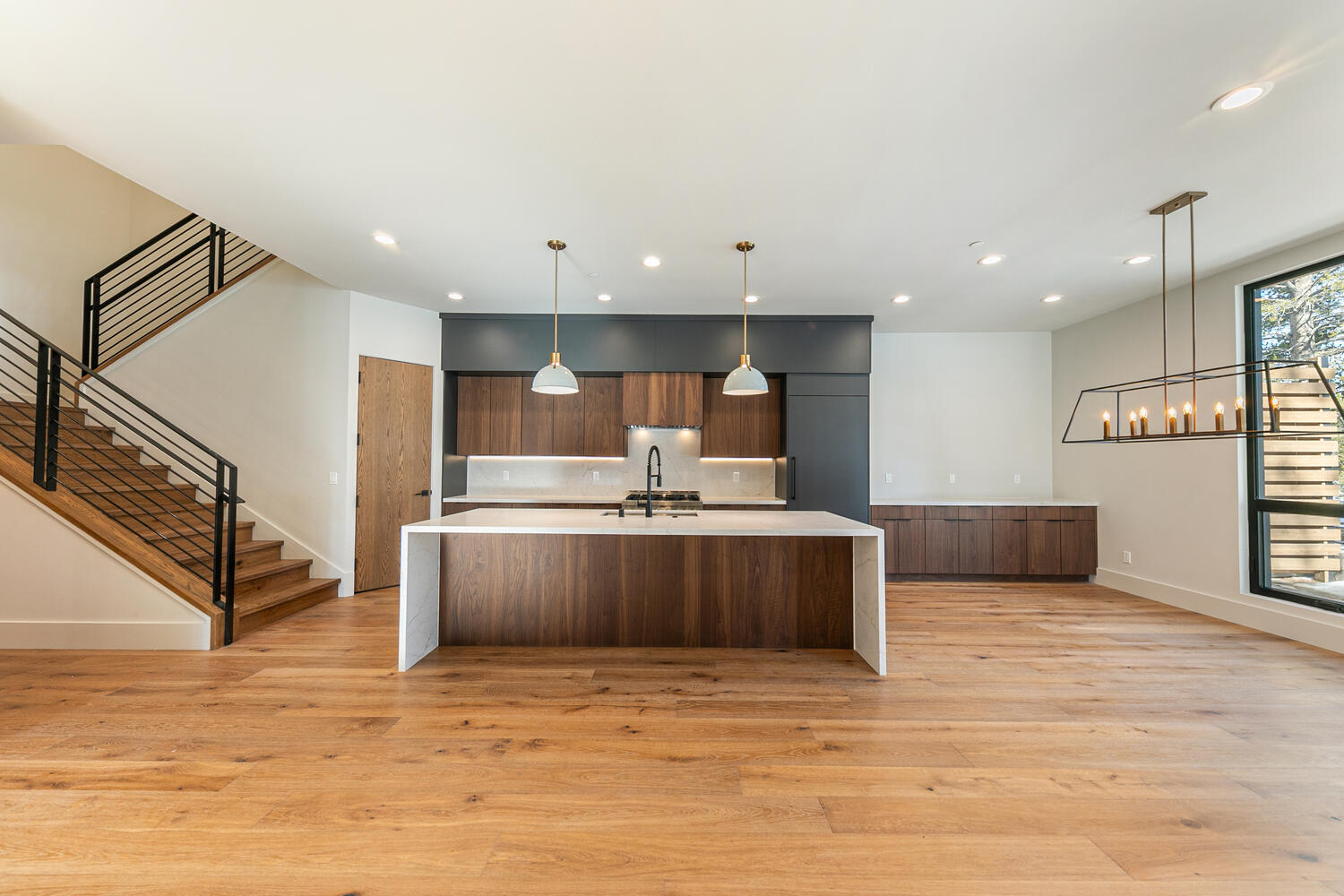 This is a modern kitchen with a large central island featuring a waterfall countertop and wooden cabinetry. The design includes dark-colored upper cabinets and accent lighting, along with sleek appliances. The kitchen also incorporates a staircase, hardwood flooring, and a dining area with a view from a large window, presenting an open and inviting living space.