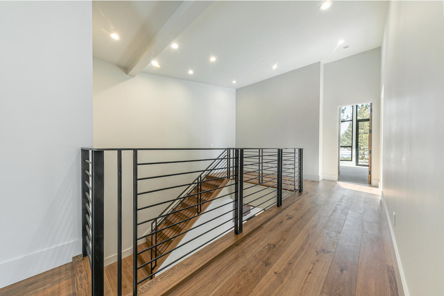 This interior shot showcases a modern hallway and staircase. The hallway features hardwood flooring and white walls, creating a bright and spacious feel. The staircase with black railing leads to a lower level, adding architectural interest to the home.