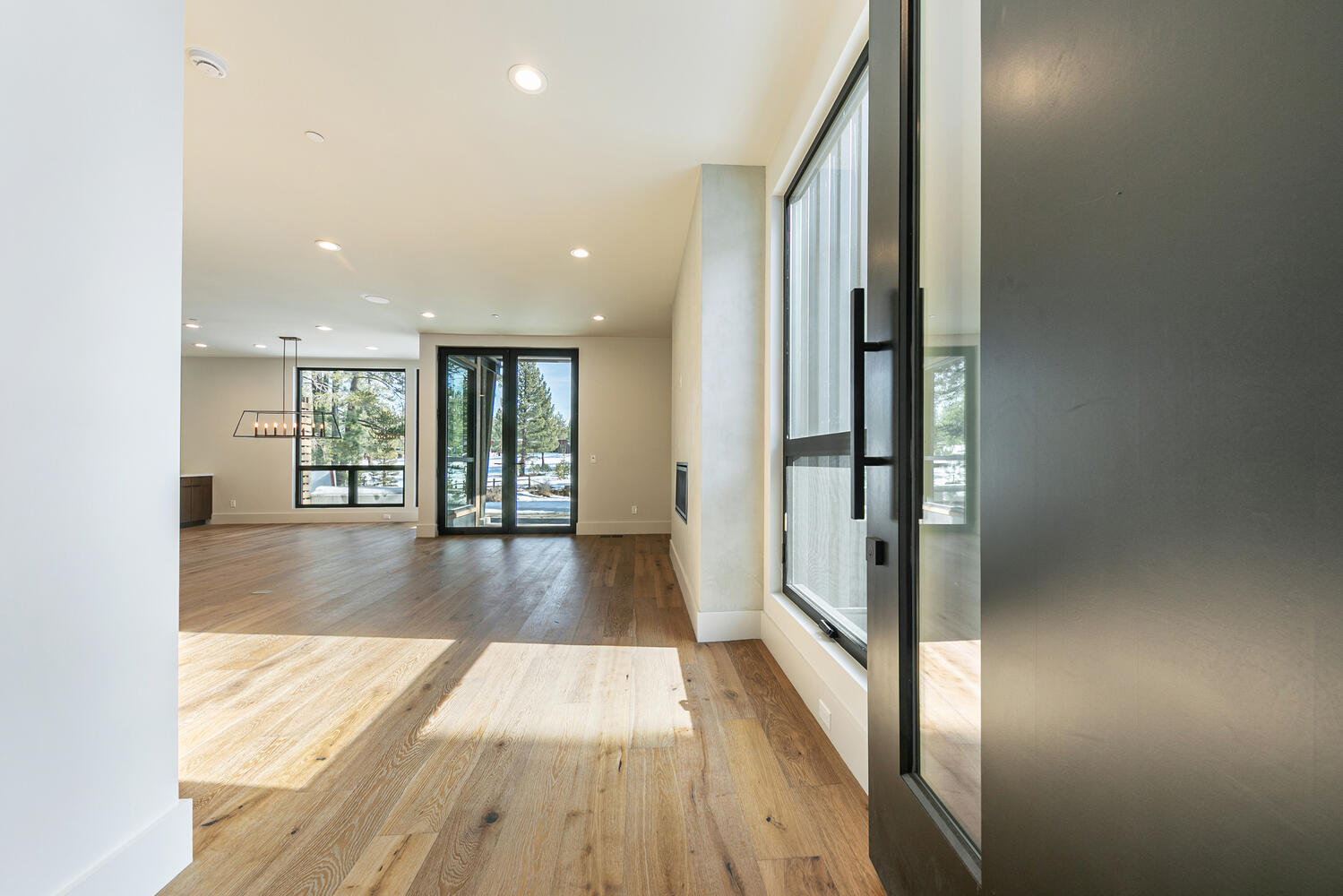 This interior shot showcases a modern living room with an open floor plan and hardwood flooring. Large black-framed windows offer ample natural light and views of the outdoors. The entrance shown features a dark door with glass panels, complementing the contemporary aesthetic of the space.