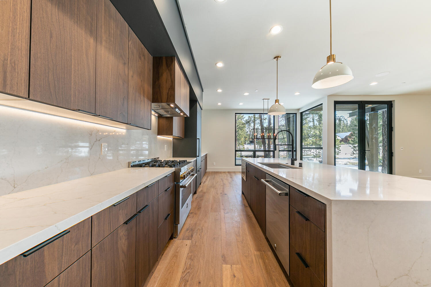 This is a modern kitchen with wooden cabinets, a white countertop, and stainless steel appliances including a range. It features track lighting underneath the upper cabinetry and pendant lighting above a large island. A hardwood floor stretches throughout the kitchen.