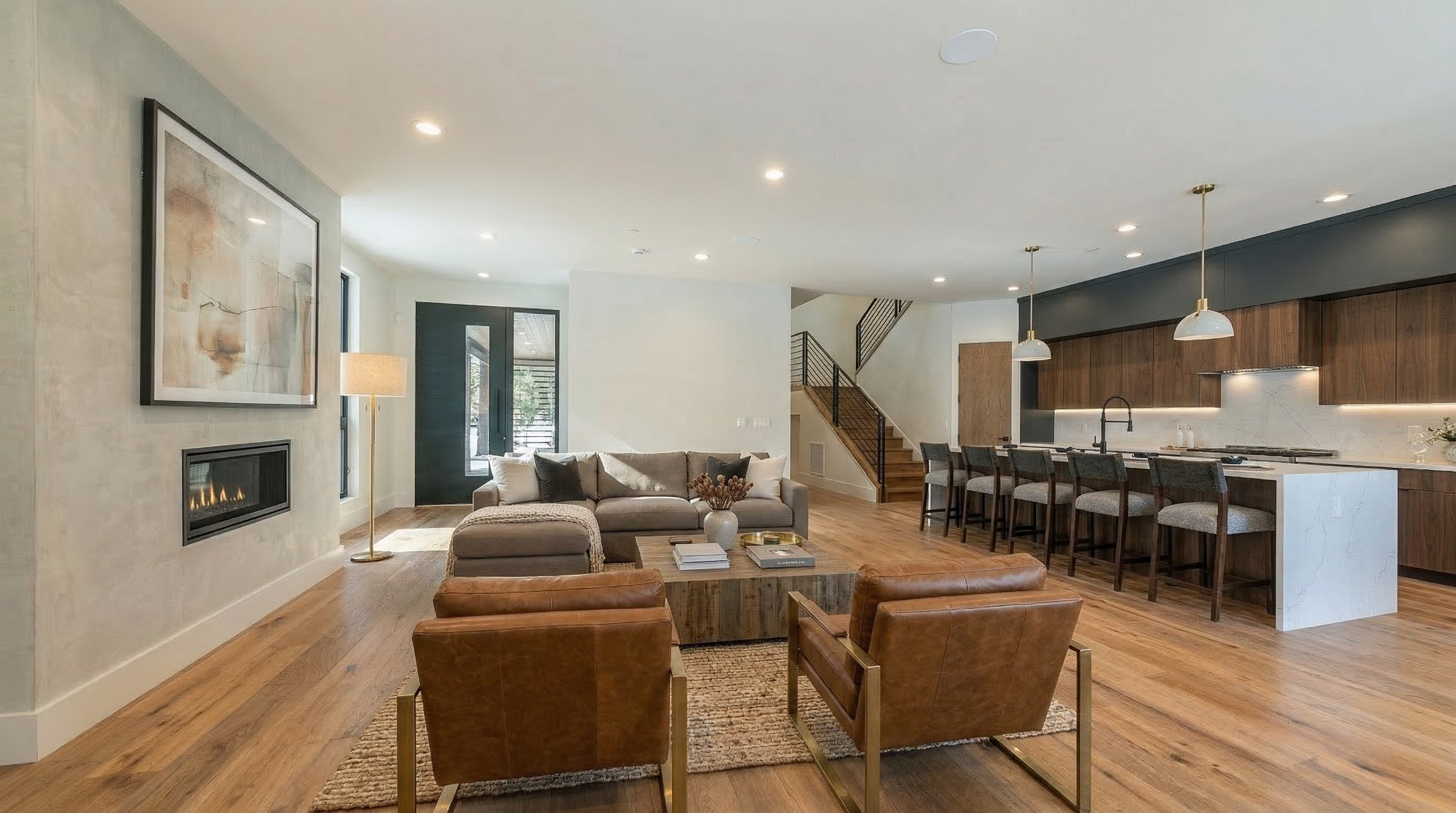 This is an interior shot of a living room. The style is modern with hardwood floors, a fireplace, and an open concept layout that flows into the kitchen. There are two chairs in the foreground, a sofa in the middle, and bar stools in the kitchen area.