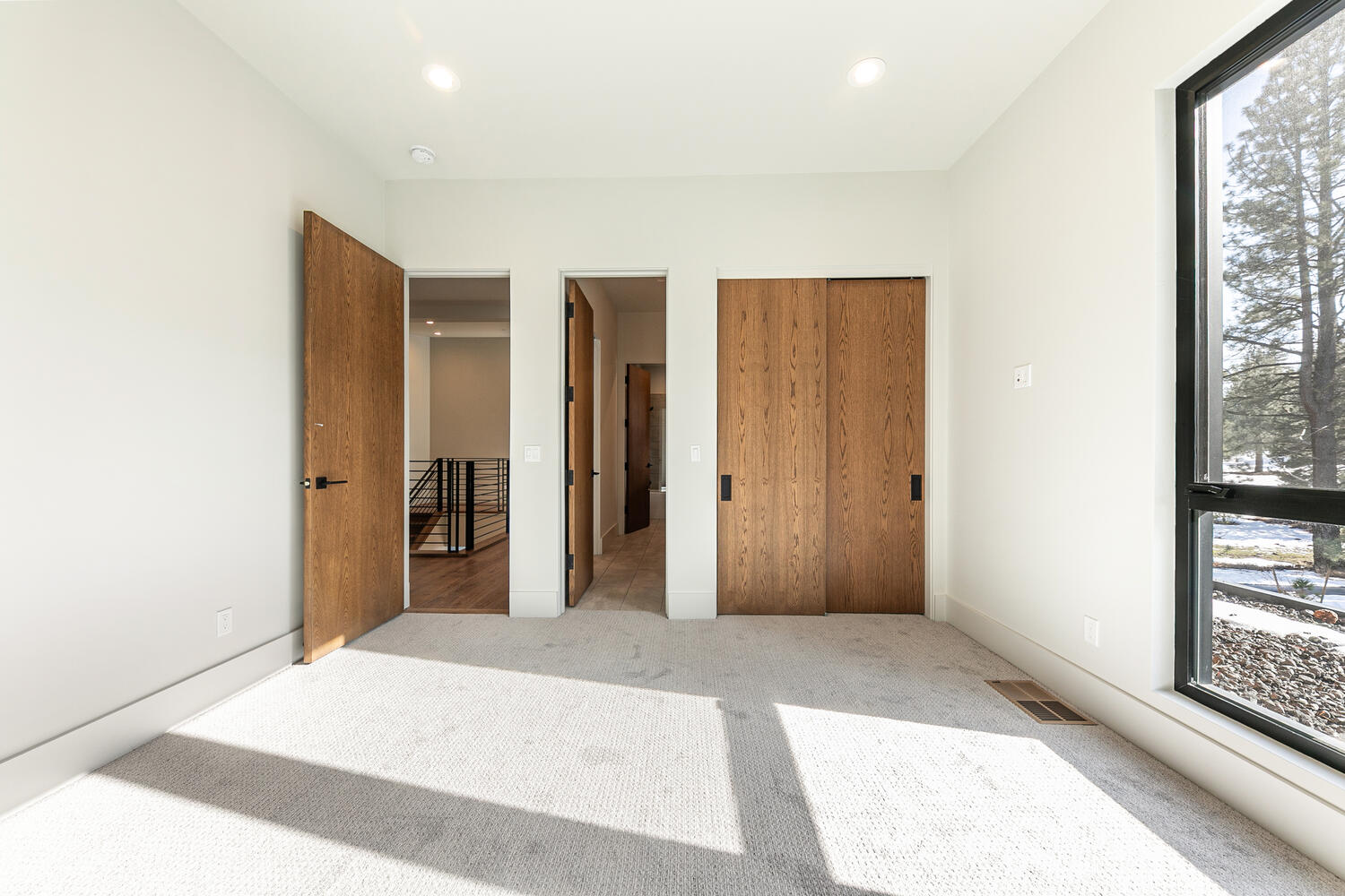 The image showcases a well-lit bedroom interior, likely a guest or secondary bedroom, featuring neutral wall colors and light-colored carpeting. The room includes wooden doors that open to reveal interior spaces, plus a large window offering natural light and a view. The overall aesthetic is modern and minimalist, creating a serene and inviting impression.