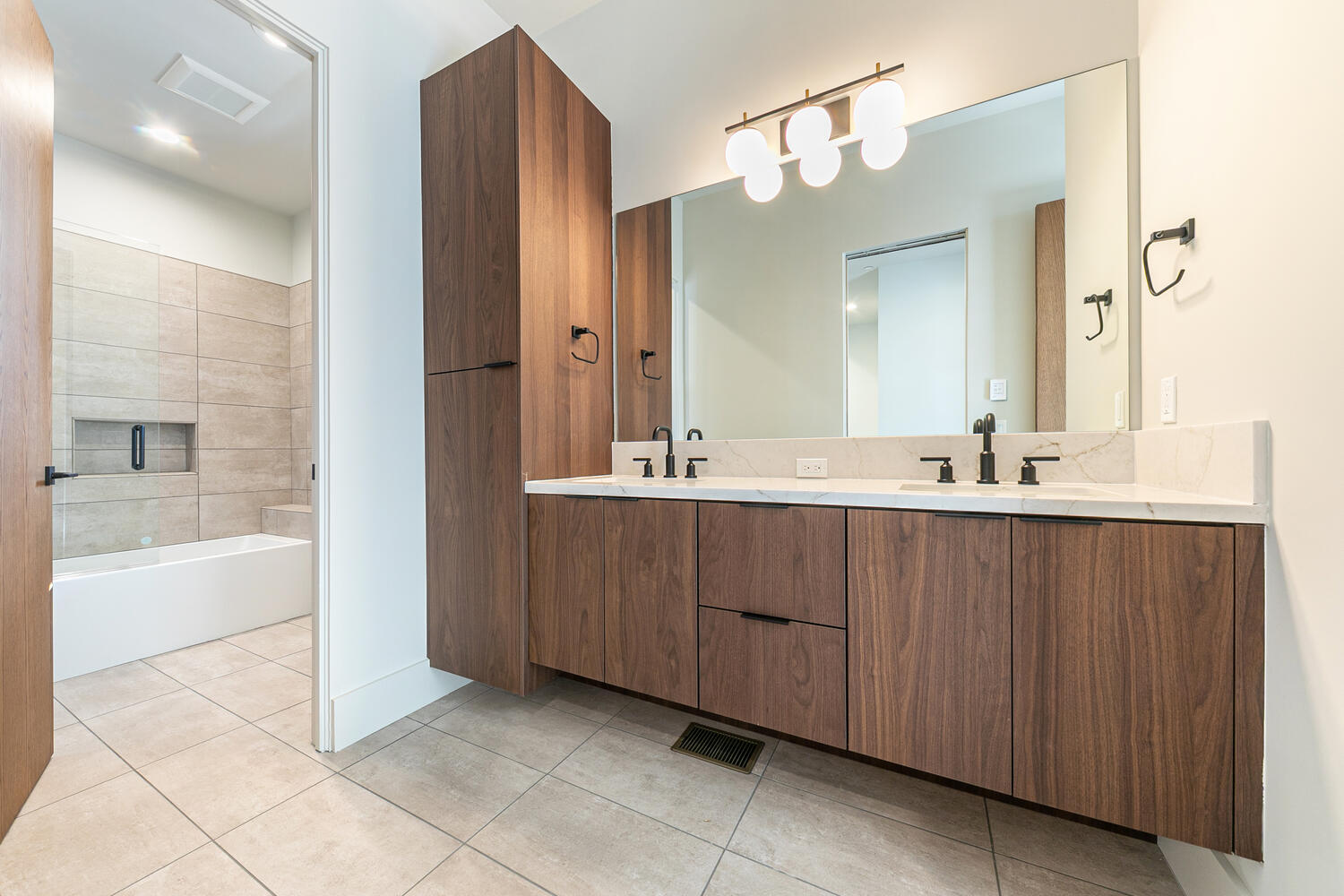 The image showcases a modern primary bathroom featuring a double vanity with what appears to be a marble countertop and dark wood cabinetry. A large mirror is centered above the vanity with a contemporary light fixture. The bathroom extends into a wet room with a bathtub and light beige tiling contributing to a clean, minimalist aesthetic.
