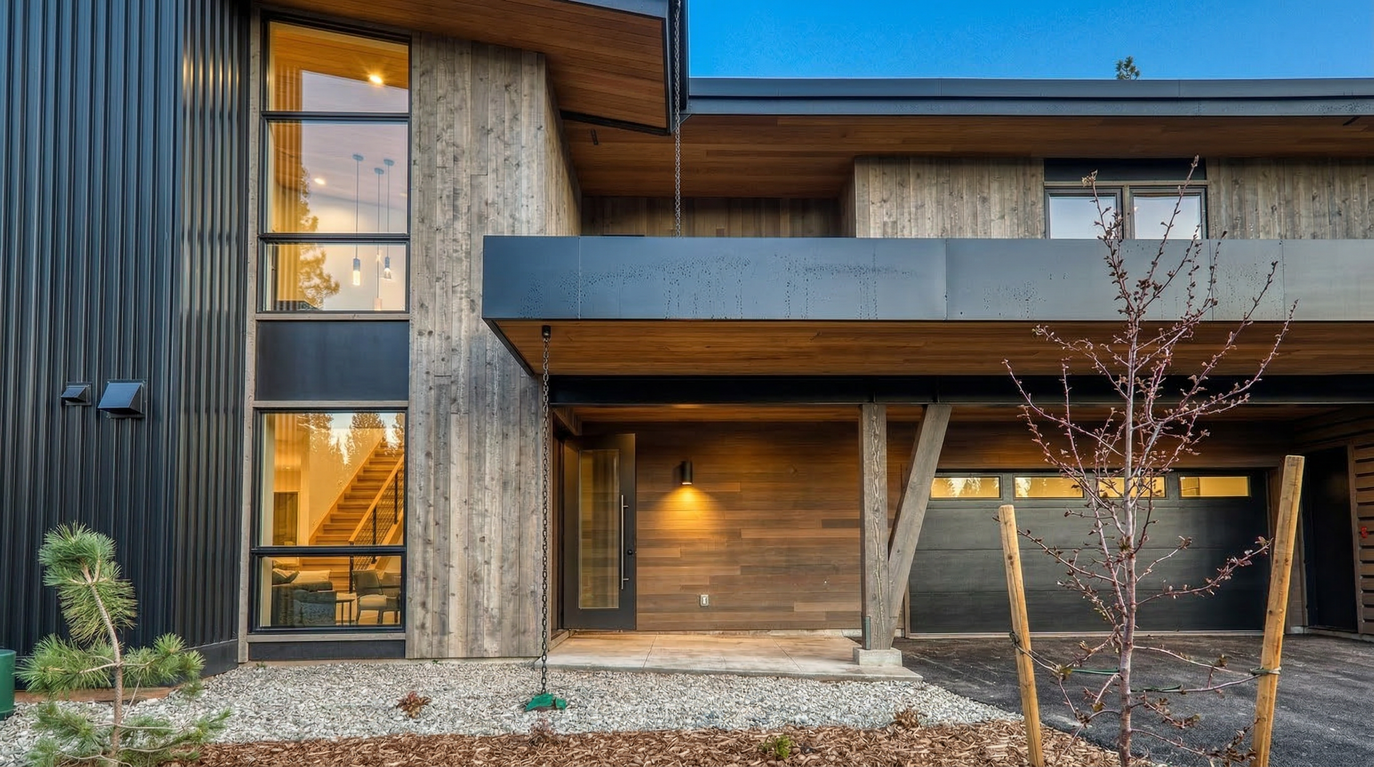 This is a front exterior view showcasing a modern house with a unique blend of materials including dark metal siding and natural wood accents. Large windows offer glimpses of the home's interior, and the entryway is covered by a cantilevered overhang providing shelter. The landscaping features a mix of gravel and wood chips, complementing the overall contemporary design.