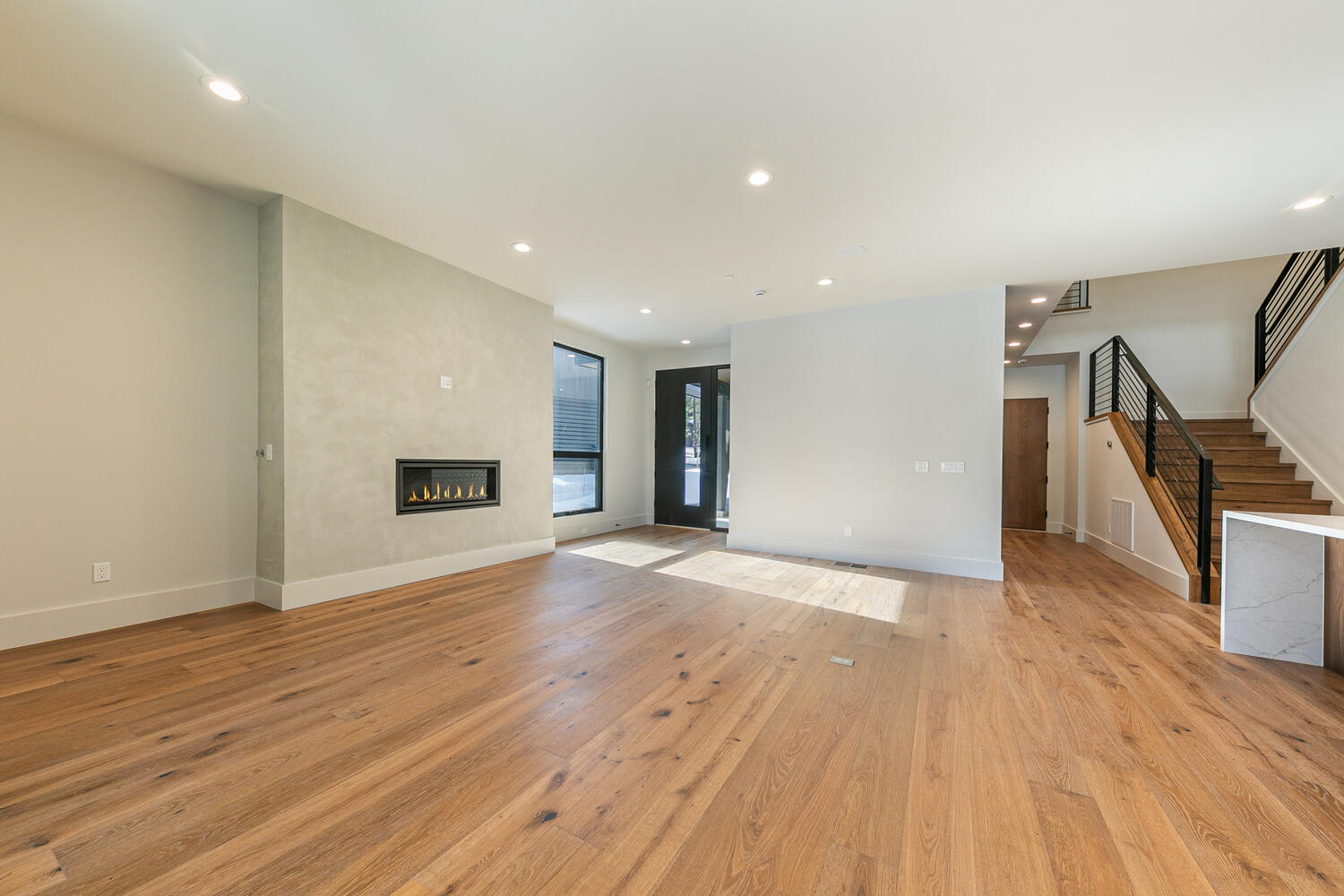 This is a bright and spacious living room with hardwood flooring. It features a modern fireplace, a large window, and an open staircase with metal railings. The room has a clean and minimalist design, perfect for modern living.