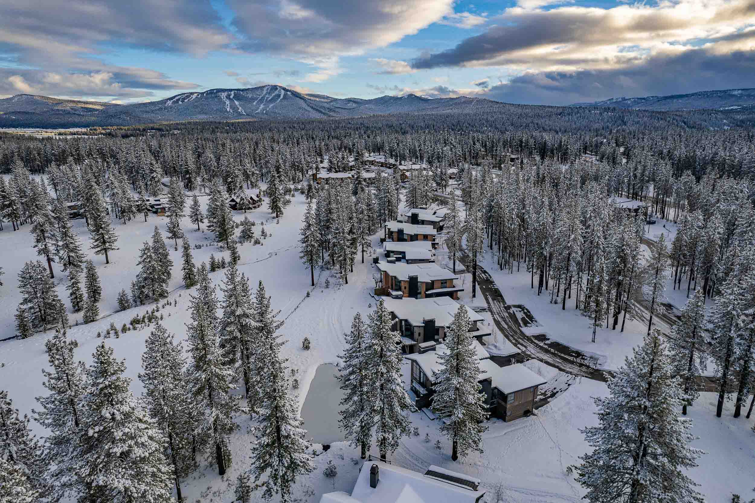 This aerial view showcases a cluster of modern homes nestled in a snow-covered forest, possibly in a mountain resort community. The architecture features clean lines and dark exterior finishes, blending harmoniously with the wintry landscape. A winding road provides access to the properties, suggesting a tranquil and secluded atmosphere.