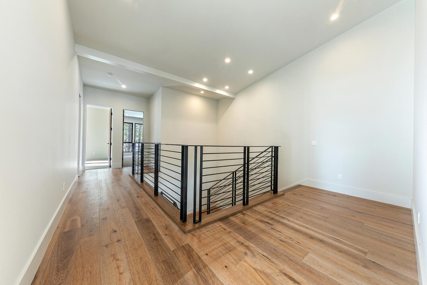 This interior shot showcases a modern hallway and staircase area. The space features light hardwood flooring, white walls, and a black metal railing. Recessed lighting brightens the area, creating a clean and contemporary atmosphere.