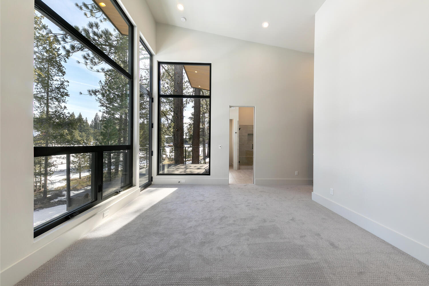 This is the primary bedroom of a modern home. The room features large format windows that let in plenty of natural light and offer a view of the surrounding landscape. The walls are painted in a neutral color, and the flooring is light gray carpet. There is an en suite bathroom connected to the room through a doorway.