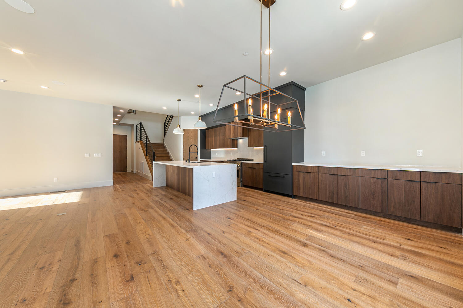 This spacious interior showcases a modern kitchen and living area featuring hardwood flooring throughout. A large kitchen island boasts a white countertop, complemented by streamlined cabinetry and a contemporary light fixture. The open floor plan enhances the airy feel of the space, making it ideal for entertaining and family gatherings.