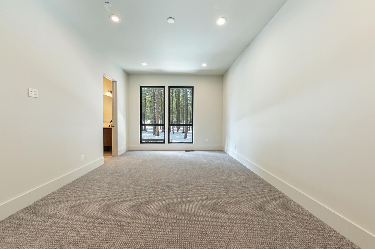 This is a bright and spacious primary bedroom with neutral tones. The room features carpet flooring, recessed lighting, and two windows providing natural light and a view of the outdoors. A door on the left suggests access to an ensuite bathroom.