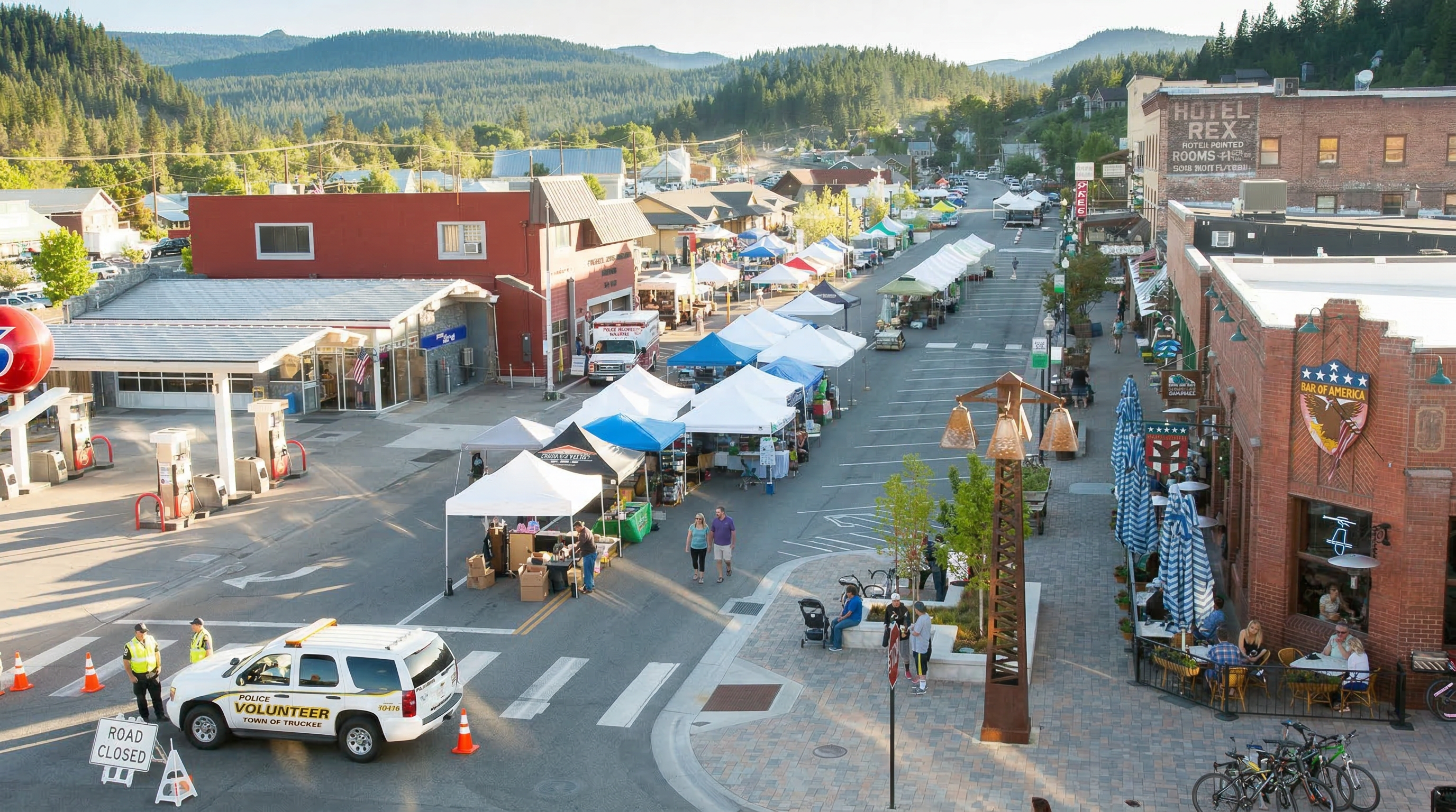 This aerial view showcases a vibrant small-town setting, featuring a street lined with vendor tents and storefronts. Notable features include a vintage gas station, the Hotel Rex, and the Bar of America, creating a charming commercial area. A police volunteer vehicle and officers suggest a community event or road closure, adding to the scene's lively atmosphere.