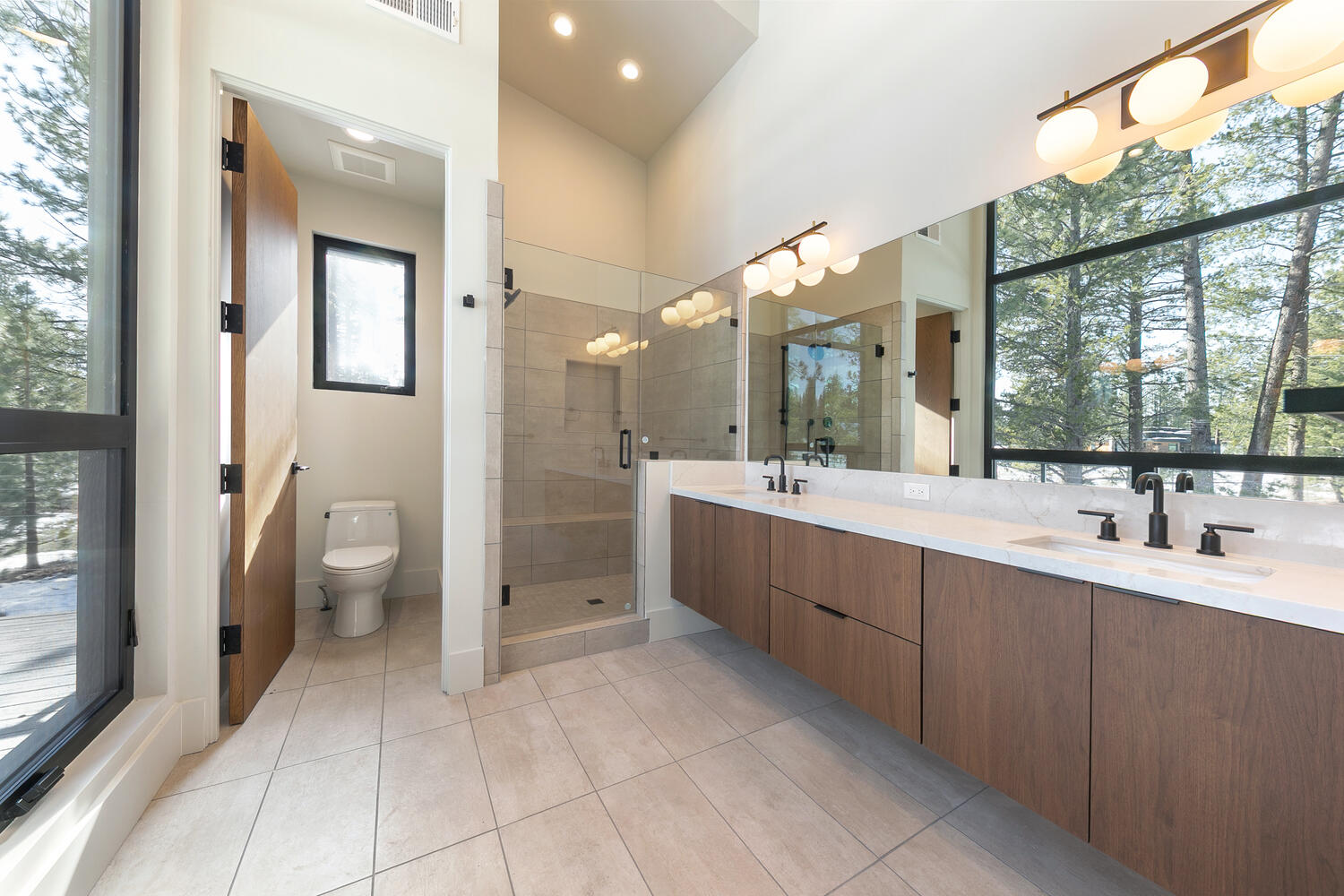 This primary bathroom boasts a modern design with a double vanity featuring wood-grain cabinetry and a bright white countertop. A large mirror reflects natural light from a nearby window, and a glass-enclosed shower is visible in the background. The neutral tile flooring complements the upscale and serene ambiance of the space.