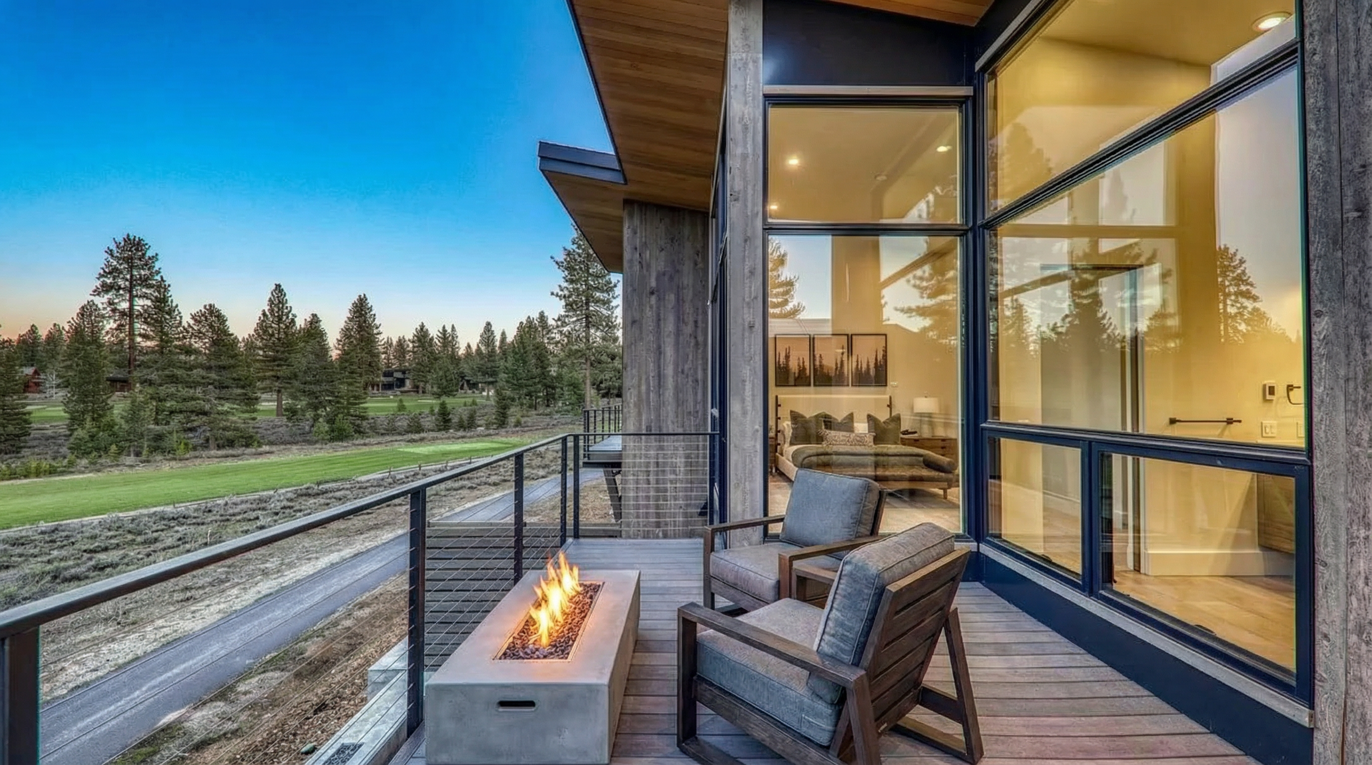 This inviting outdoor living space features a modern deck with a fire pit, perfect for enjoying cool evenings. Two comfortable armchairs are arranged to take in the view. The space overlooks a lush, green landscape, possibly a golf course, and large windows provide peeks into the interior, suggesting a seamless indoor-outdoor lifestyle.