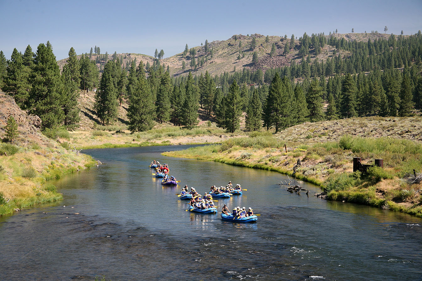 This photo showcases a community amenity, specifically a scenic river used for recreational activities such as rafting. The image highlights the community's access to nature and outdoor adventures, with multiple rafts filled with people enjoying the waterway. Lush green trees and a hill with scattered trees line the banks of the river.