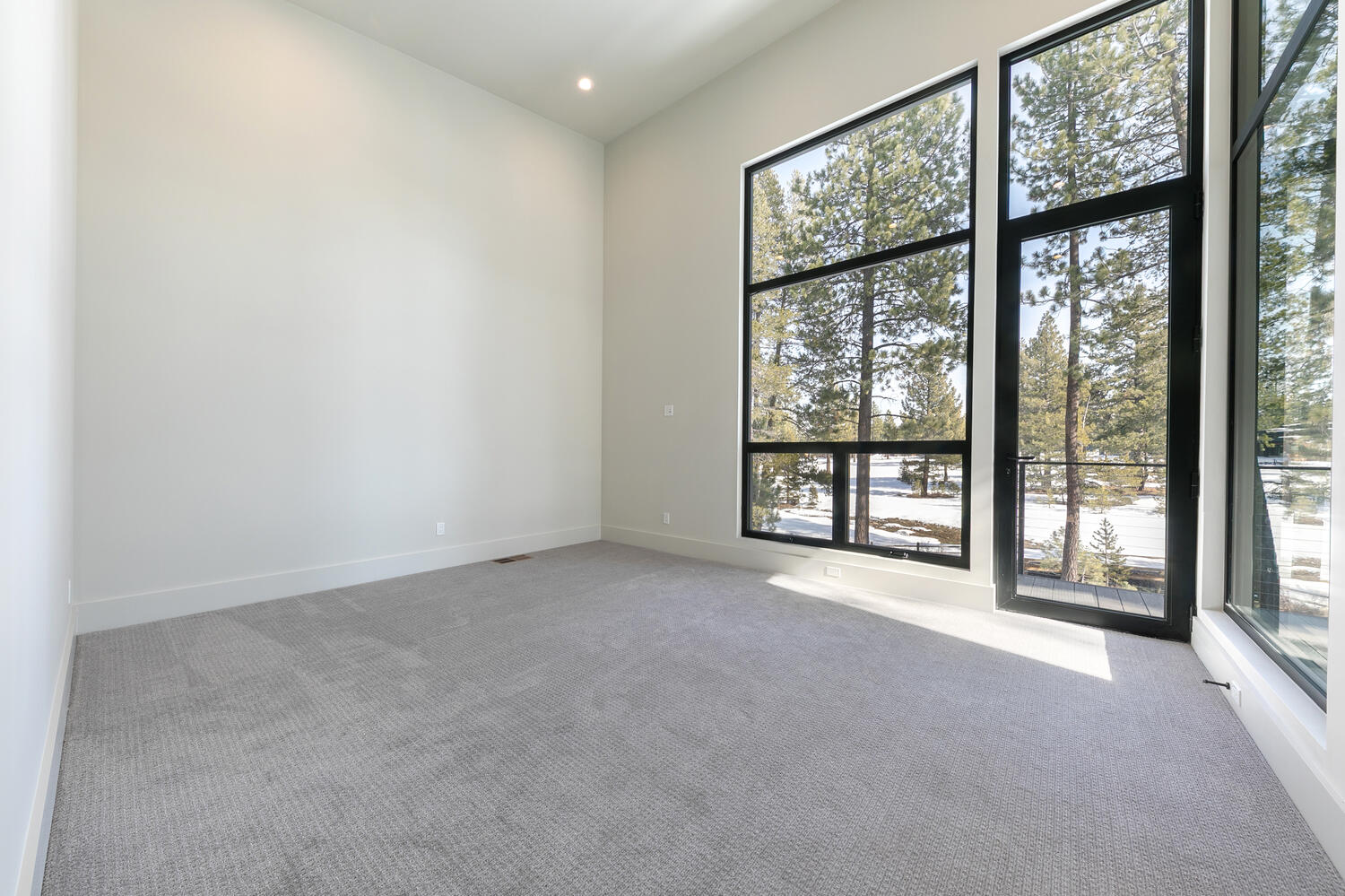 This is a bright and spacious primary bedroom with modern design. Large windows with black frames offer ample natural light and views of the outdoors. The room features neutral-colored walls, light gray carpeting, and minimalist trim, providing a blank canvas for the future homeowner.