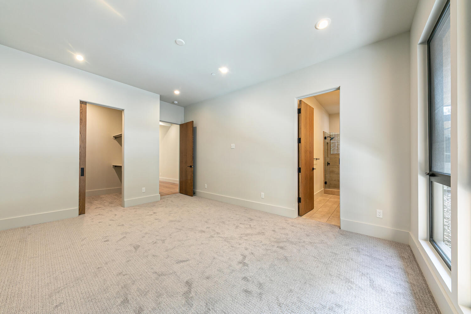 This is a spacious primary bedroom featuring neutral carpeting and paint tones. The room has two doorways leading to a walk-in closet and an ensuite bathroom, offering convenient access. The large window provides ample natural light and enhances the room's open feel, making it an appealing setting for rest and relaxation.