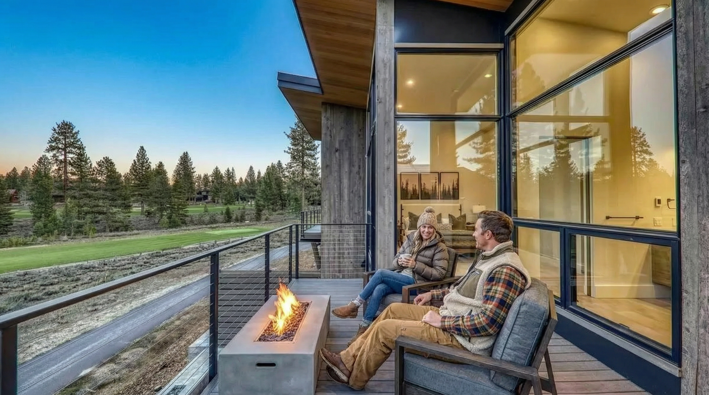 This image showcases a modern patio or balcony featuring an outdoor fireplace around which a couple sits, implying a cozy and relaxing atmosphere. The space overlooks a scenic view of trees and what appears to be a golf course. The design incorporates modern architectural elements with large windows facing into the home.