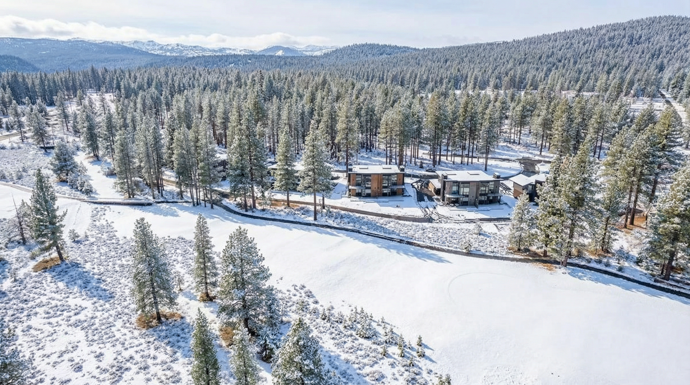 This aerial shot displays beautifully constructed modern homes nestled in a snow-covered forest. The properties feature contemporary designs with clean lines and neutral tones, blending harmoniously with the natural surroundings. The snowy landscape enhances the sense of tranquility and privacy, making it an attractive location.